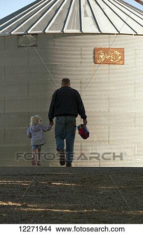 Picture - A farmer and his young daughter walk hand-in-hand through his farm yard with a grain bin in the background, near Sioux City; Iowa, United States of America. Fotosearch