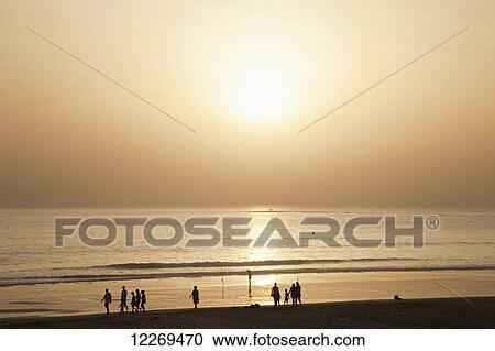 Stock Image - A glowing sun sets in a yellow sky reflecting in the tranquil ocean with silhouettes of people on the beach; Ciclana de la Frontera, Andalusia, Spain. Fotosearch