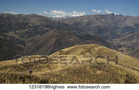 Stock Photograph - A hiker makes his way through the foothills of the Andes Mountains with the snowcapped peaks in the background of the Peruvian landscape; Cusco, Peru. Fotosearch