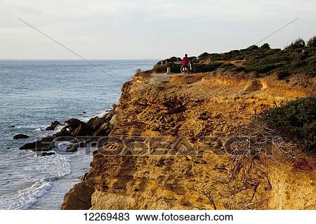 Stock Image - A male cyclist stands on the cliff ledge looking out to the ocean; Conil de la Frontera, Andalusia, Spain. Fotosearch