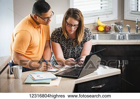 Stock Photograph - A man and woman standing in the kitchen working together with a calculator and computer; Regina, Saskatchewan, Canada. Fotosearch