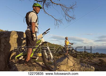 Stock Photography - A man and woman standing with their bicycles on a rocky ledge overlooking a view of the landscape; Tarifa, Cadiz, Andalusia, Spain. Fotosearch