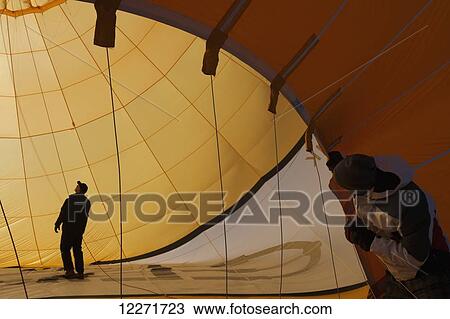 Stock Image - A man looks on as his colleague checks ropes inside a hot air balloon as it lies on the ground being inflated; Filzmoos, Austria. Fotosearch