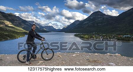 A man sits on a bicycle at the water's edge with the Rocky mountains and a lake in the background, Waterton Lakes National Park; Alberta, Canada View Large Photo Image Stock Photograph - A man sits on a bicycle at the water's edge with the Rocky mountains and a lake in the background, Waterton Lakes National Park; Alberta, Canada. Fotosearch