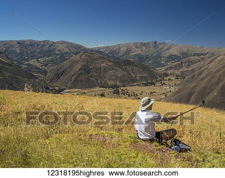 Stock Photograph - A man takes a selfie with his selfie-stick while resting during a hike in the foothills of the Andes Mountains; Cusco, Peru. Fotosearch