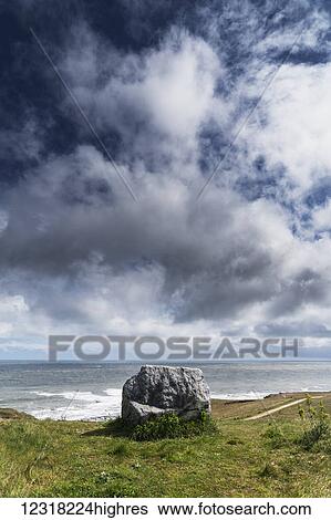 Stock Photograph - A rock sits on the grassy shore with a view of a rail along the ocean under a cloudy sky; South Shields, Tyne and Wear, England. Fotosearch