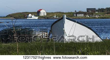 Stock Photograph - A rowboat on the grass shore with buildings and houses along the coast, Cape Breton Island; Nova Scotia, Canada. Fotosearch