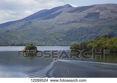 Picture - A small boat floats through tranquil water of the Lakes of Killarney surrounded by mountains; Killarney, County Kerry, Ireland. Fotosearch