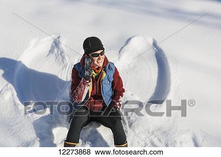 Stock Photo - A smiling young woman wearing a hat and gloves talks on her cell phone after making a snow angel in a snowbank, Winter, Southcentral Alaska, USA. Fotosearch