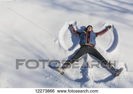 Stock Photograph - A smiling young woman wearing a hat and gloves makes a snow angel in a snowbank, Winter, Southcentral Alaska, USA. Fotosearch