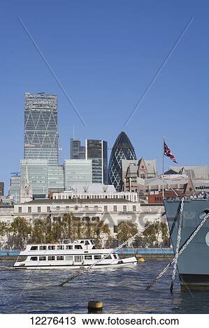 Stock Image - A tourist boat passes the prow of the warship HMS Belfast, docked on the River Thames and part of the Imperial War Museum, with skyscrapers in the City of London, including The Gherkin by Norman Foster; London, England. Fotosearch