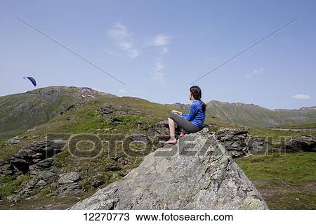 Stock Image - A tween girl sitting on a boulder watching para gliders sail across the sky at Hatcher Pass, Talkeetna Mountains, Southcentral Alaska, Summer. Fotosearch