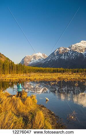 Stock Photo - A woman and her dog are standing by a pond on with the Chugach Mountains reflecting in a pond in the background, Eagle River Valley, Chugach State Park, Southcentral Alaska. . Fotosearch