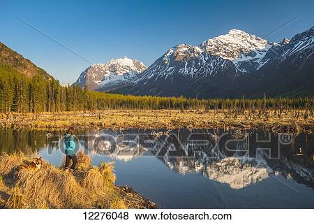 Stock Photo - A woman and her dog are standing by a pond on with the Chugach Mountains reflecting in a pond in the background, Eagle River Valley, Chugach State Park, Southcentral Alaska.. Fotosearch