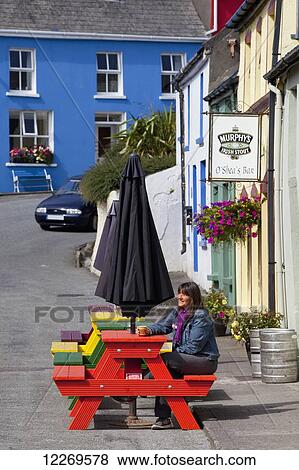 Stock Photo - A woman sits outside a bar on a colourful picnic table; Eyeries, County Cork, Ireland. Fotosearch