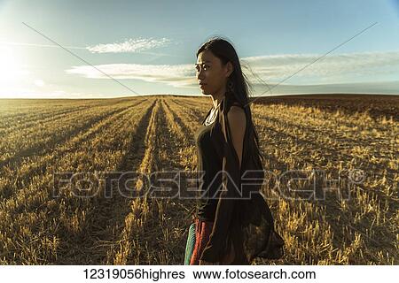 Stock Photograph - A young Asian woman walking in a golden wheat field; Madrid, Spain. Fotosearch