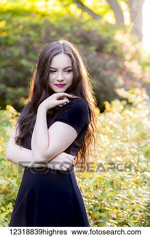 Stock Photograph - A young millennial teenage girl in a fashionable black dress stands in a thoughtful pose in a sunny park setting; Vancouver, British Columbia, Canada. Fotosearch
