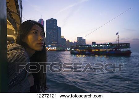 Stock Image - A young woman at the waterfront at sunset with a a boat and skyline in the background, Kowloon; Hong Kong, China. Fotosearch