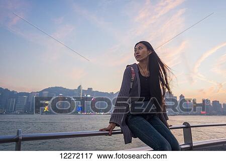 Stock Photo - A young woman at the waterfront sitting on the railing at sunset with the skyline in the background, Kowloon; Hong Kong, China. Fotosearch