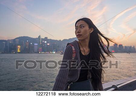 Stock Photo - A young woman at the waterfront sitting on the railing at sunset with the skyline in the background, Kowloon; Hong Kong, China. Fotosearch