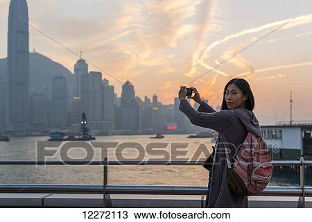 Stock Image - A young woman at the waterfront taking pictures of the skyline at sunset, Kowloon; Hong Kong, China. Fotosearch
