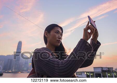 A young woman at the waterfront taking a picture at sunset with the skyline in the background, Kowloon; Hong Kong, China View Large Photo Image Stock Photo - A young woman at the waterfront taking a picture at sunset with the skyline in the background, Kowloon; Hong Kong, China. Fotosearch