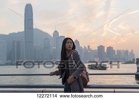 Picture - A young woman at the waterfront with a view of the skyline at sunset, Kowloon; Hong Kong, China. Fotosearch