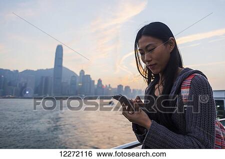 A young woman checks her cell phone at the waterfront with a view of the skyline at sunset, Kowloon; Hong Kong, China View Large Photo Image Stock Photograph - A young woman checks her cell phone at the waterfront with a view of the skyline at sunset, Kowloon; Hong Kong, China. Fotosearch