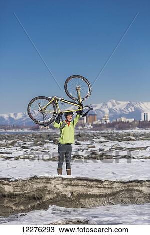 Stock Image - A young woman holds her bicycle over her head in triumph while standing on ice next to the Tony Knowles Coastal Trail, Anchorage, Southcentral Alaska, USA.. Fotosearch