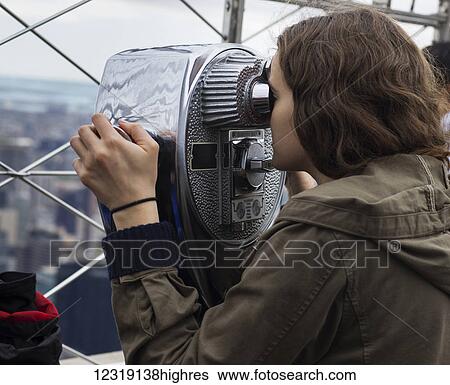 Stock Photograph - A young woman looks through binoculars on the observation deck of the Empire State building to a view of the cityscape of New York City; New York City, New York, United States of America. Fotosearch