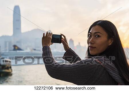 A young woman photographs buildings across the water, Kowloon; Hong Kong, China View Large Photo Image Stock Photograph - A young woman photographs buildings across the water, Kowloon; Hong Kong, China. Fotosearch
