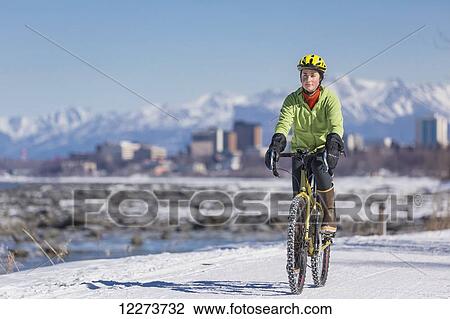 Stock Image - A Young Woman rides a studded tire bicycle down the Tony Knowles Coastal Trail, Anchorage, Southcentral Alaska, USA.. Fotosearch