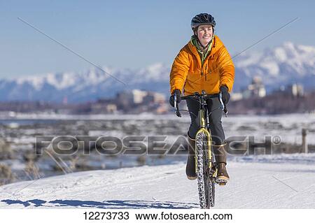 Stock Image - A Young Woman rides a studded tire bicycle down the Tony Knowles Coastal Trail, Anchorage, Southcentral Alaska, USA.. Fotosearch