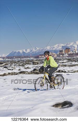 Picture - A young woman riding her bicycle on ice and snow next to the Tony Knowles Coastal Trail, Anchorage, Southcentral Alaska, USA.. Fotosearch