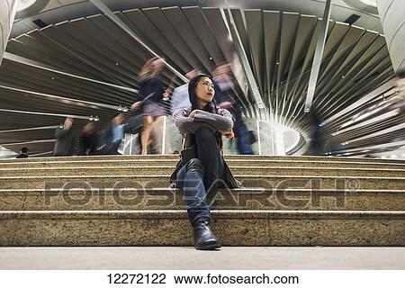 Stock Image - A young woman sitting on steps with a modern building and pedestrians in the background, Kowloon; Hong Kong, China. Fotosearch