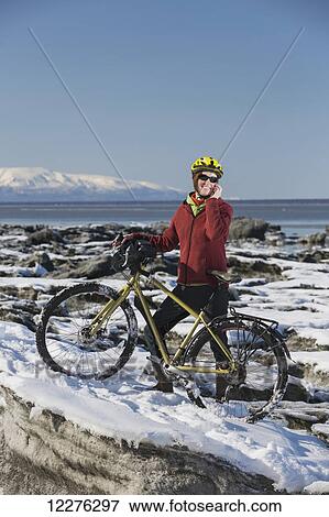Stock Photo - A young woman stands with her bicycle on ice while talking on a cell phone next to the Tony Knowles Coastal Trail, Anchorage, Southcentral Alaska, USA.. Fotosearch