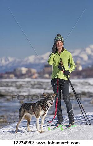 Stock Image - A young woman takes a break from skijoring with her Alaska Husky sled dog to talk on her phone, Tony Knowles Coastal Trail, Anchorage, Southcentral Alaska, USA.. Fotosearch