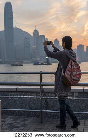 Stock Image - A young woman takes a picture with her camera of the harbour and Hong Kong skyline at sunset, Kowloon; Hong Kong, China. Fotosearch