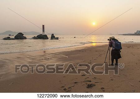 A young woman taking a picture on the beach at sunset; Xiamen, Fujian Province, China View Large Photo Image Stock Image - A young woman taking a picture on the beach at sunset; Xiamen, Fujian Province, China. Fotosearch