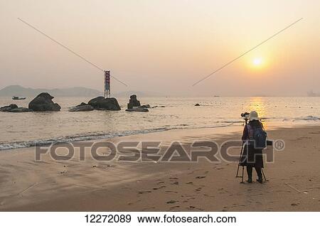 A young woman taking a picture on the beach at sunset; Xiamen, Fujian Province, China View Large Photo Image Stock Photo - A young woman taking a picture on the beach at sunset; Xiamen, Fujian Province, China. Fotosearch