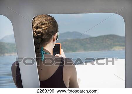 Stock Photograph - A young woman with cell phone sits at the front of a boat with a view of the ocean and coastline; Ixtapa-Zihuatanejo, Guerrero, Mexico. Fotosearch