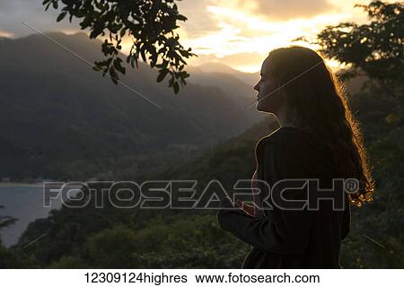 Stock Photograph - A young woman with long hair backlit by the glowing sunlight at sunset stands looking out over the ocean; Yalepa, Jalisco, Mexico. Fotosearch