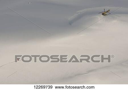 Stock Photo - Aerial view of a snowboarder walking towards a helicopter perched on the snow in mountains above Haines, Southeast Alaska, USA. Fotosearch
