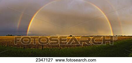 Stock Photo - Agriculture - A brilliant double rainbow forms over a farmstead and field after a Spring storm. The previous year's corn stubble is still present in the field prior to planting / Central Iowa, USA.. Fotosearch
