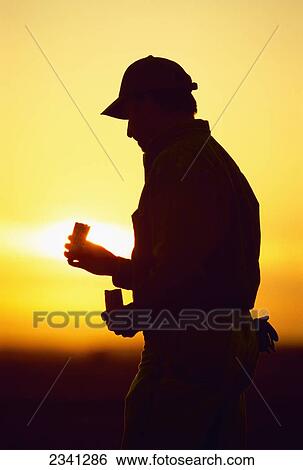 Stock Photograph - Agriculture - A farmer silhouetted by the setting sun inspects an ear of grain corn during harvest season / Central Iowa, USA.. Fotosearch