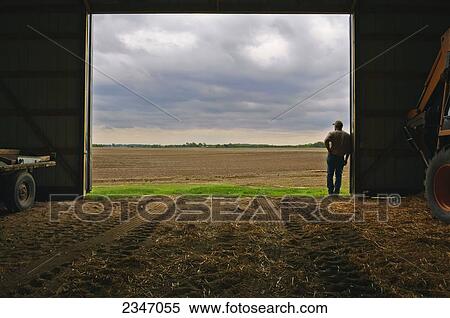 Stock Photography - Agriculture - A farmer standing at the entrance of his equipment barn looking out at approaching storm clouds and his prepared field prior to Spring planting / Iowa, USA.. Fotosearch