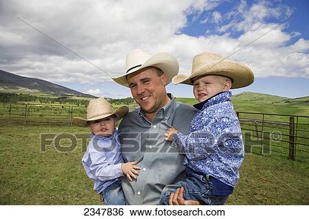 Stock Photograph - Agriculture - A Montana cowboy poses in a corral while holding his two young sons / near Stanford, Montana, USA.. Fotosearch