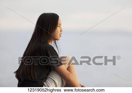 Stock Photograph - An Asian teenage girl sits looking out over the water; British Columbia, Canada. Fotosearch