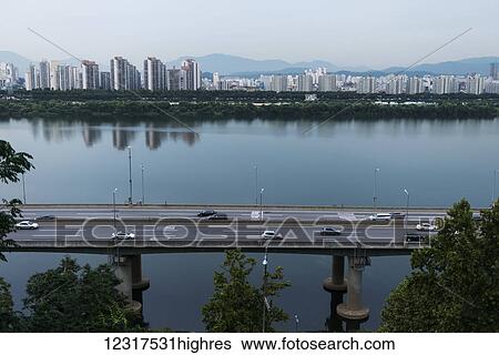 Stock Photograph - Buildings with a mountain range in the distance and a road going over water in the foreground; Seoul, South Korea. Fotosearch