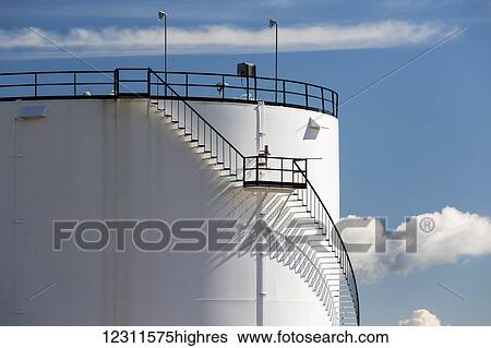 Stock Photograph - Close up of a section of a large white storage tank with black iron stairs casting a shadow on the tank and blue sky and clouds; Alberta, Canada. Fotosearch